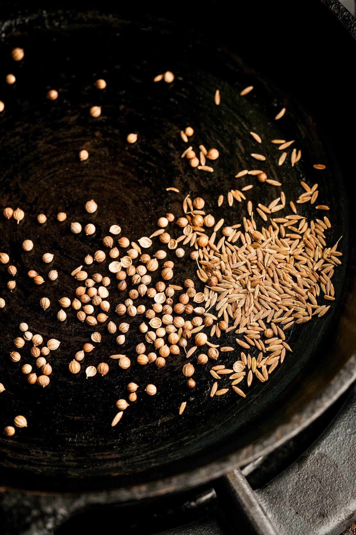 Whole coriander seeds and cumin seeds are toasting in a black skillet on a stovetop, with the seeds spread out in separate small piles.