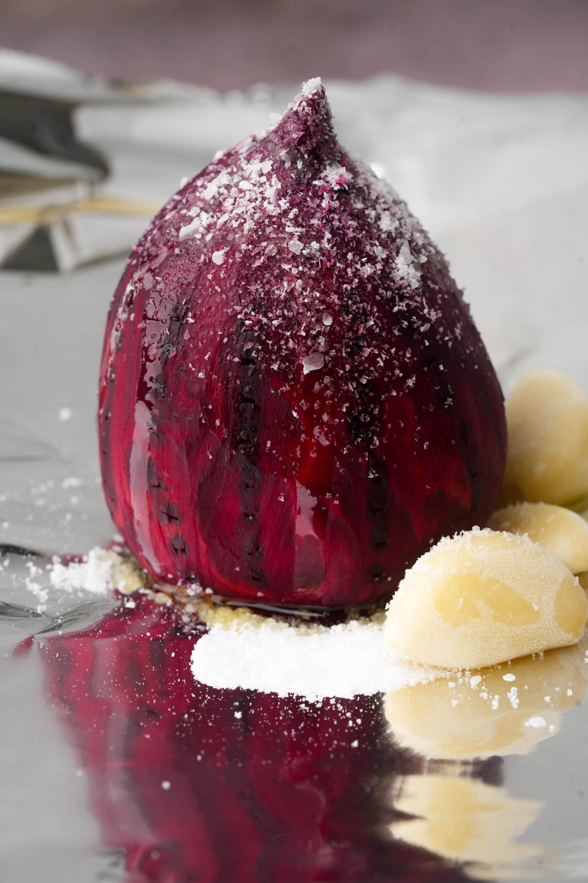 A whole red beet with a pointed top sits on foil, sprinkled with coarse salt, next to peeled garlic cloves and more salt.