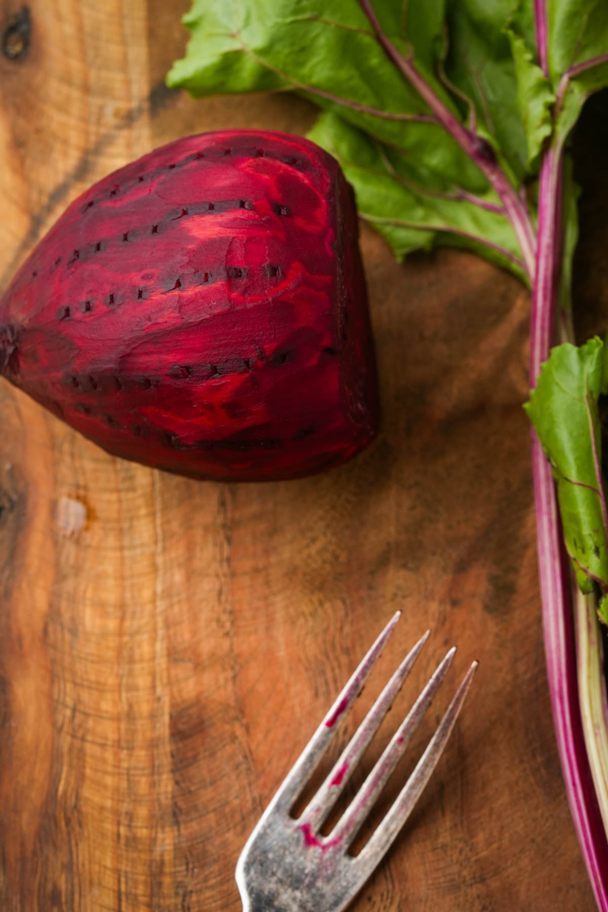 A half-peeled beet with leafy stems rests on a rustic wooden surface next to a metal fork with traces of beet juice on its tines.