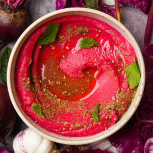 A bowl of pink beet hummus garnished with mint leaves, olive oil, and spices, surrounded by beetroot, garlic, and leafy greens on a gray surface.