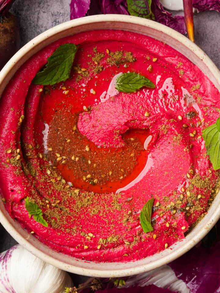 A bowl of pink beet hummus garnished with mint leaves, olive oil, and spices, surrounded by beetroot, garlic, and leafy greens on a gray surface.