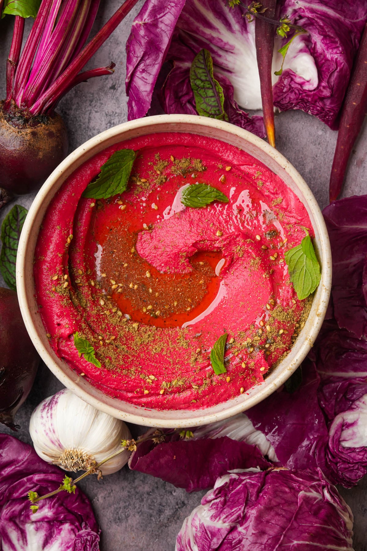 A bowl of pink beet hummus topped with olive oil, herbs, and spices, surrounded by radicchio leaves, garlic bulbs, and fresh beets on a gray surface.