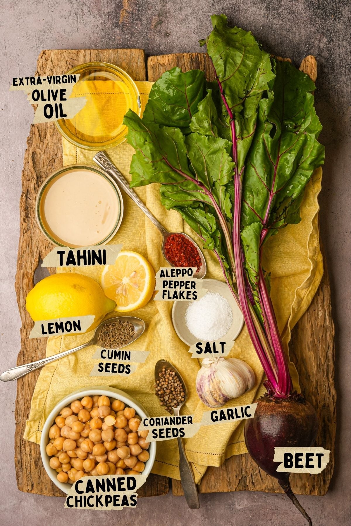 A wooden board with a yellow cloth holds labeled ingredients for a beet hummus recipe including a beet with greens, garlic, salt, red pepper flakes, cumin seeds, coriander seeds, lemon halves, tahini, olive oil, and a bowl of canned chickpeas.