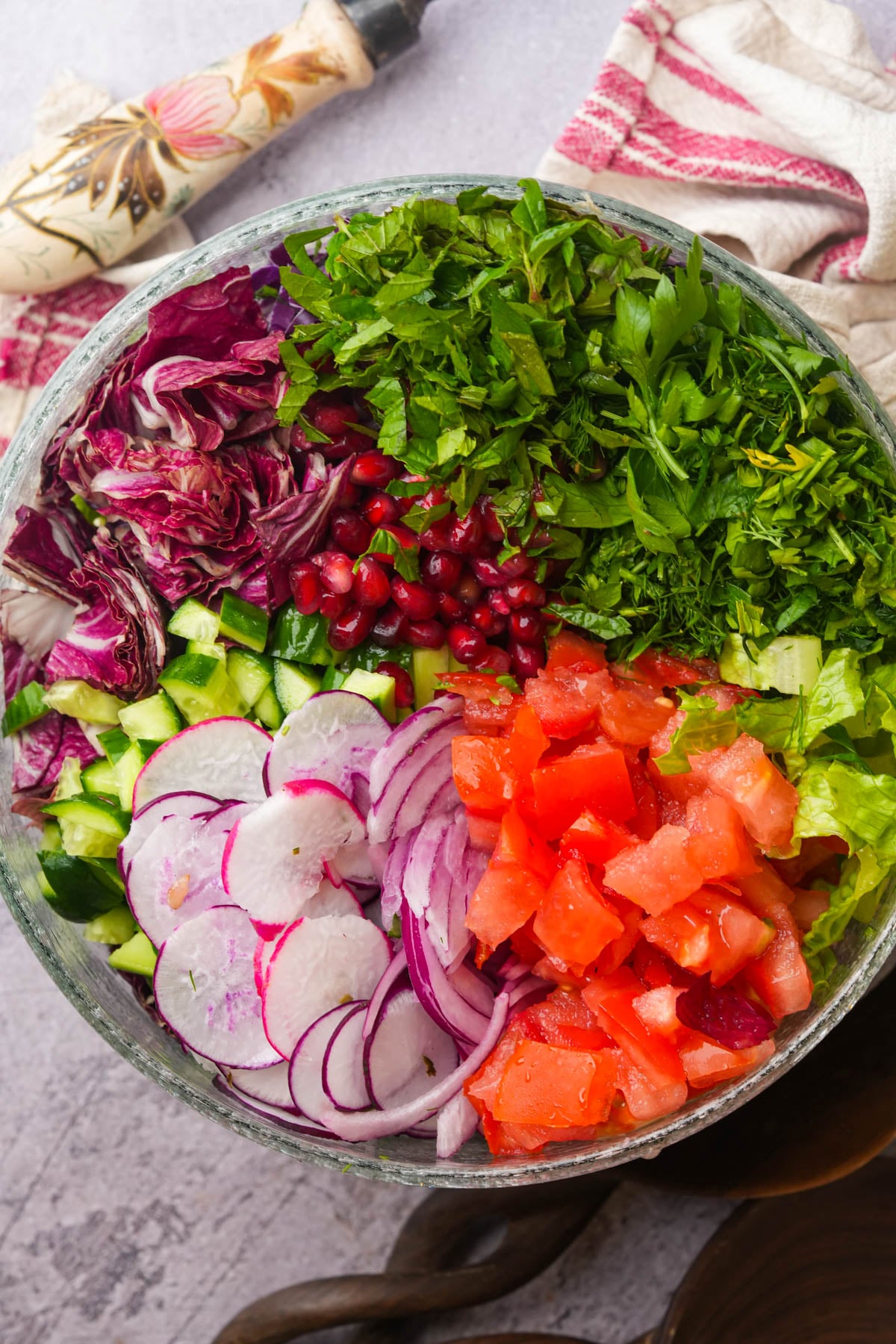 A glass bowl filled with chopped tomatoes, cucumbers, radishes, red onions, lettuce, pomegranate seeds, parsley, and radicchio, arranged in colorful sections. A towel and utensil are nearby on the table.