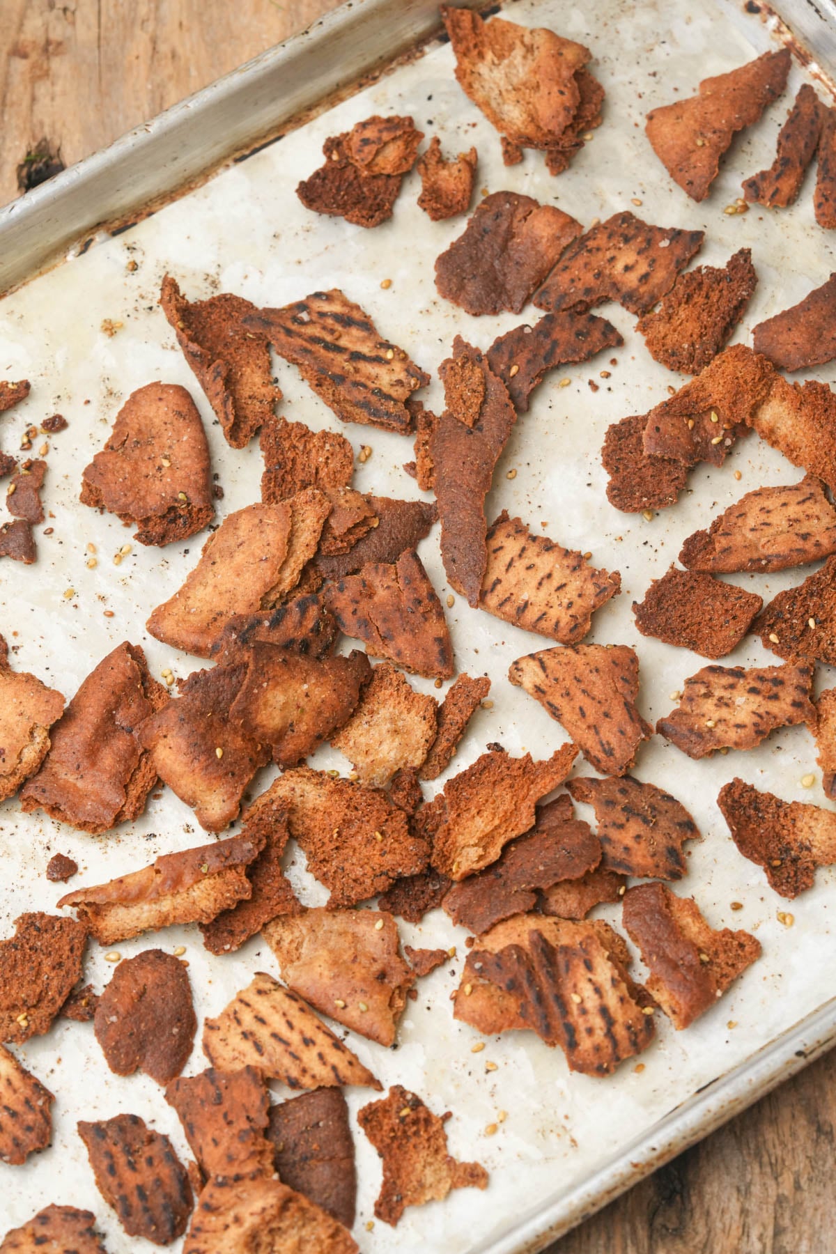 Baked pita chips scattered on a parchment-lined baking sheet, showing uneven browning and charring. The tray rests on a rustic wooden surface.