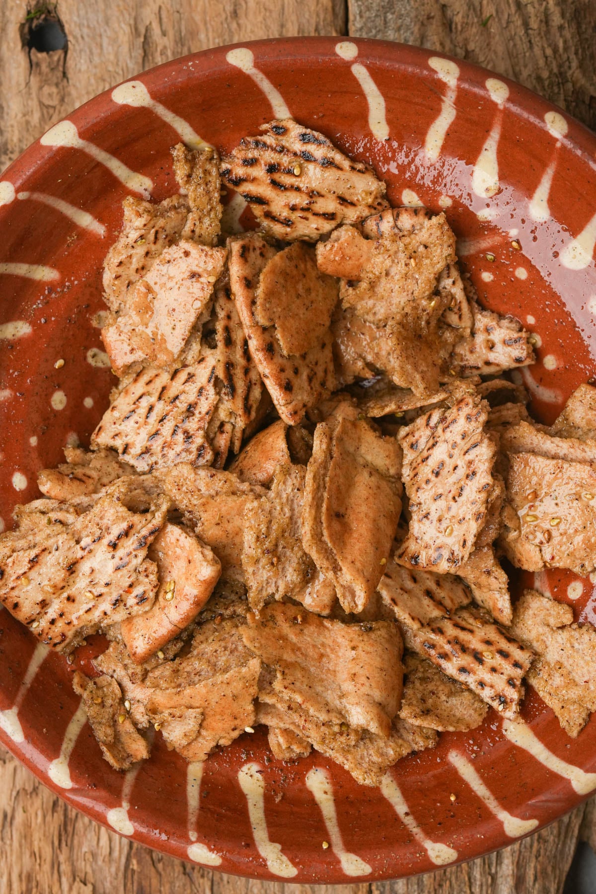 A terracotta bowl filled with small, broken pita pieces coated in spices and olive oil, sitting on a rustic wooden surface.