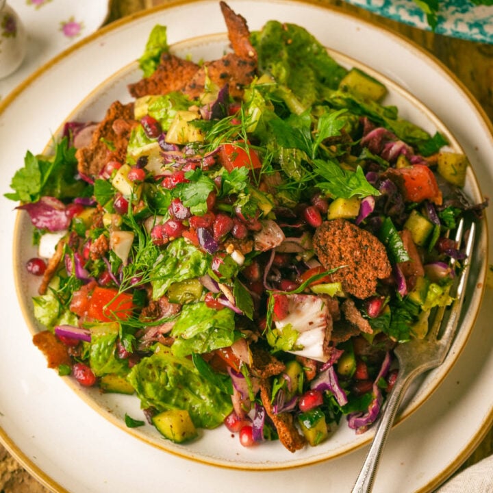 A colorful fattoush salad with lettuce, pomegranate seeds, chopped vegetables, herbs, and crispy pita pieces on a white plate with a fork.