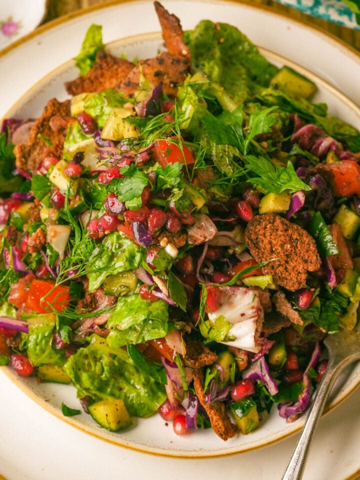 A colorful fattoush salad with lettuce, pomegranate seeds, chopped vegetables, herbs, and crispy pita pieces on a white plate with a fork.