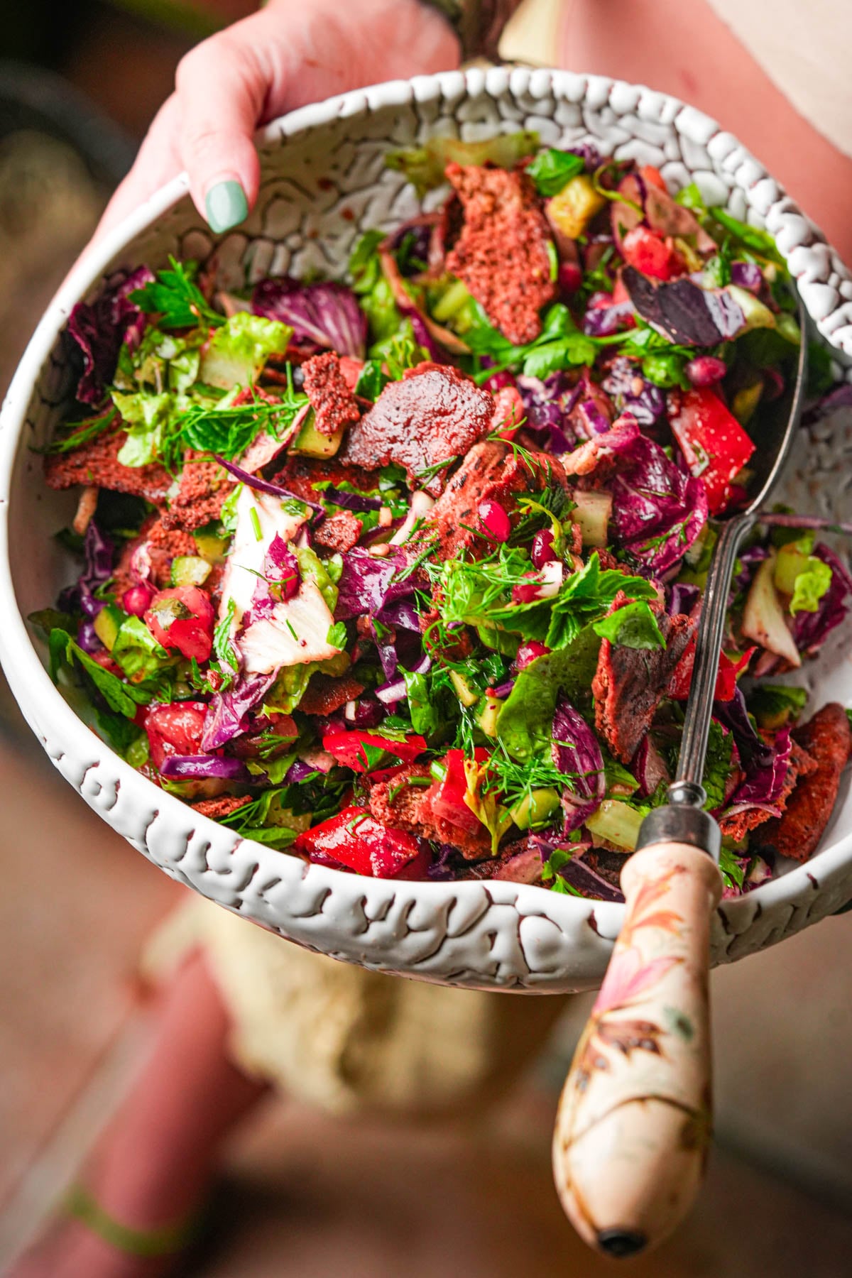 A hand holds a ceramic bowl filled with colorful fattoush salad, featuring leafy greens, tomatoes, pita chips, and herbs; a decorative spoon rests inside.