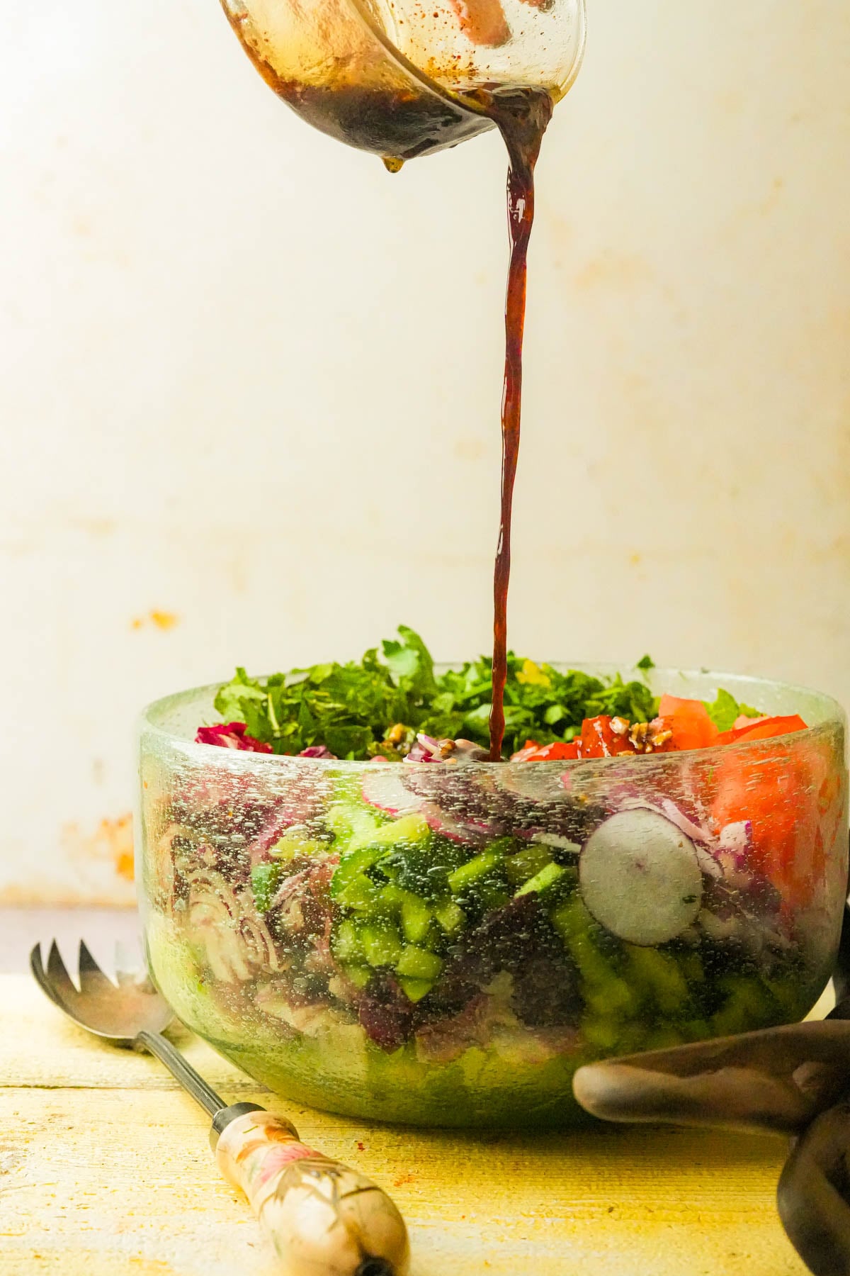 A glass bowl of fattoush including tomatoes and greens, with dark dressing being poured on top. A serving fork and spoon are placed beside the bowl on a yellow surface.