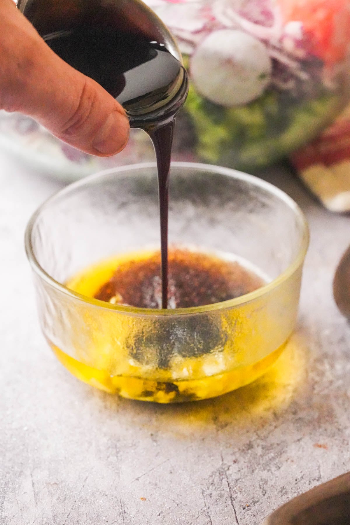 A hand pours dark pomegranate molasses into a glass bowl with olive oil and minced garlic, preparing fattoush salad dressing; fresh salad is visible in the background.