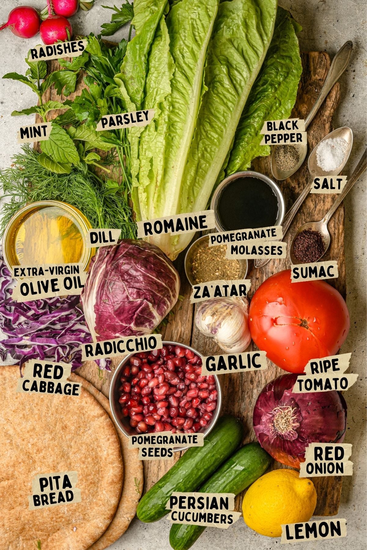 Top view of various fresh ingredients labeled for a fattoush recipe including Romaine lettuce, parsley, dill, mint, radicchio, red cabbage, tomatoes, red onion, cucumbers, lemon, pita bread, and spices on a rustic surface.