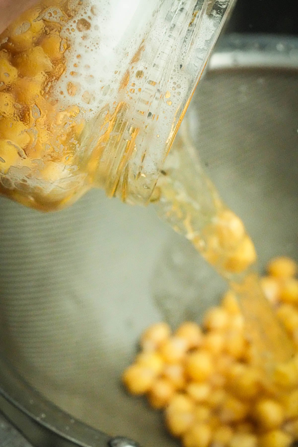 Chickpeas being poured from a glass jar into a metal strainer, with foamy liquid draining away.