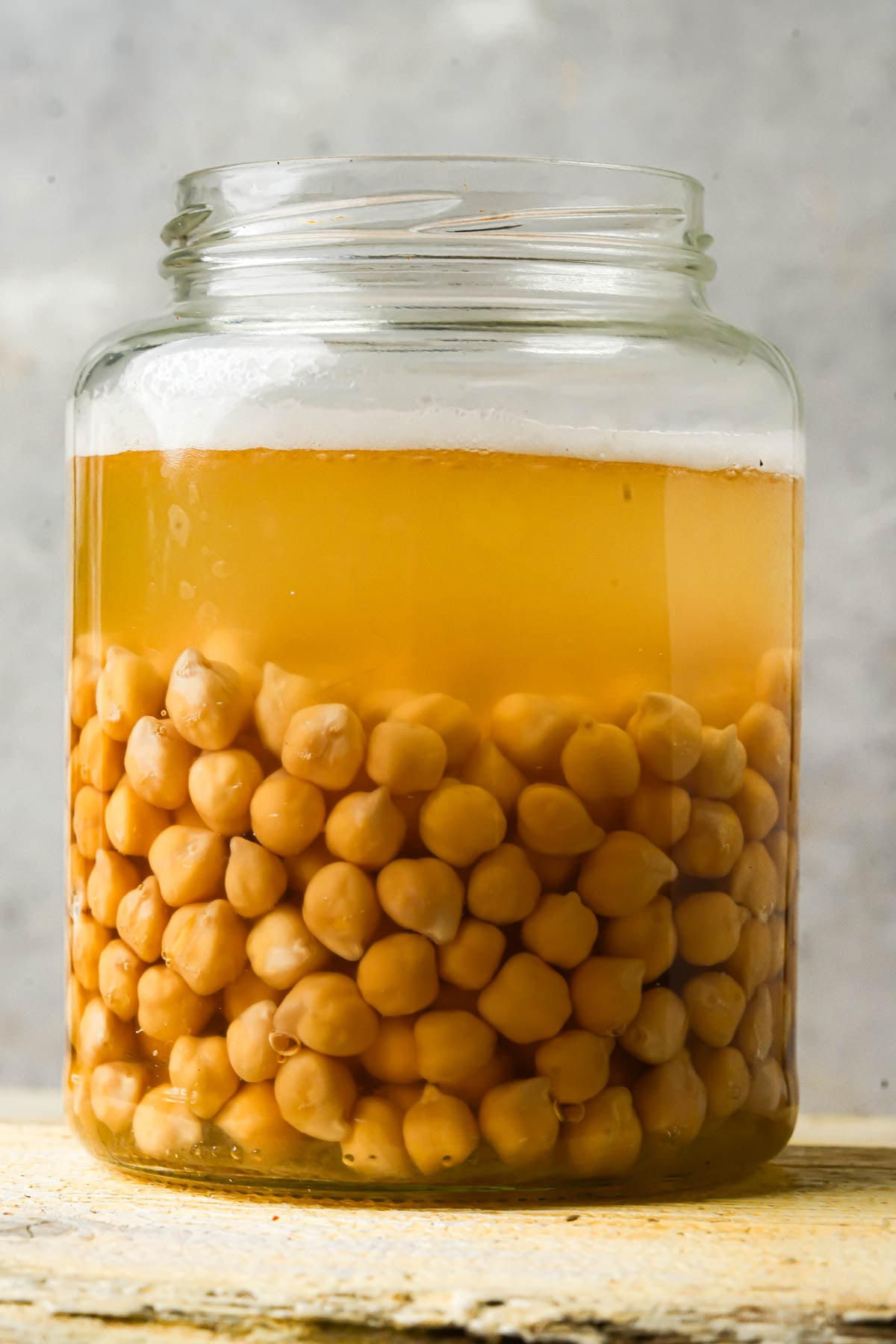 A clear glass jar filled with chickpeas yellowed liquid, with the chickpeas settled at the bottom and the water covering them, set against a neutral background.