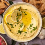 A bowl of creamy lemon hummus topped with olive oil, lemon slices, chopped parsley, and black pepper, surrounded by pita bread, lemon wedges, and a garlic bulb.