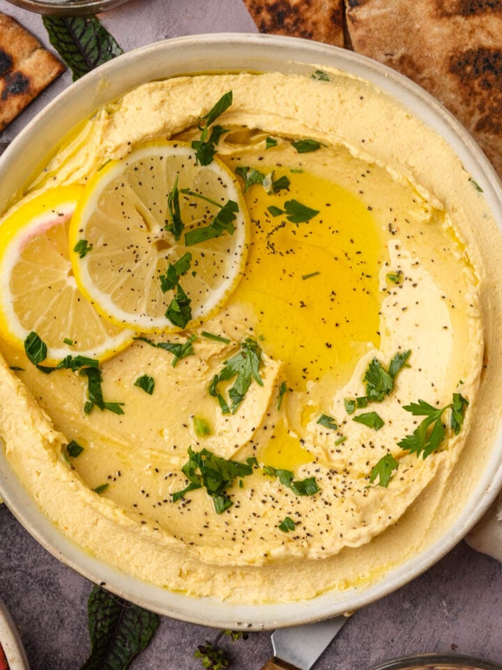 A bowl of creamy lemon hummus topped with olive oil, lemon slices, chopped parsley, and black pepper, surrounded by pita bread, lemon wedges, and a garlic bulb.