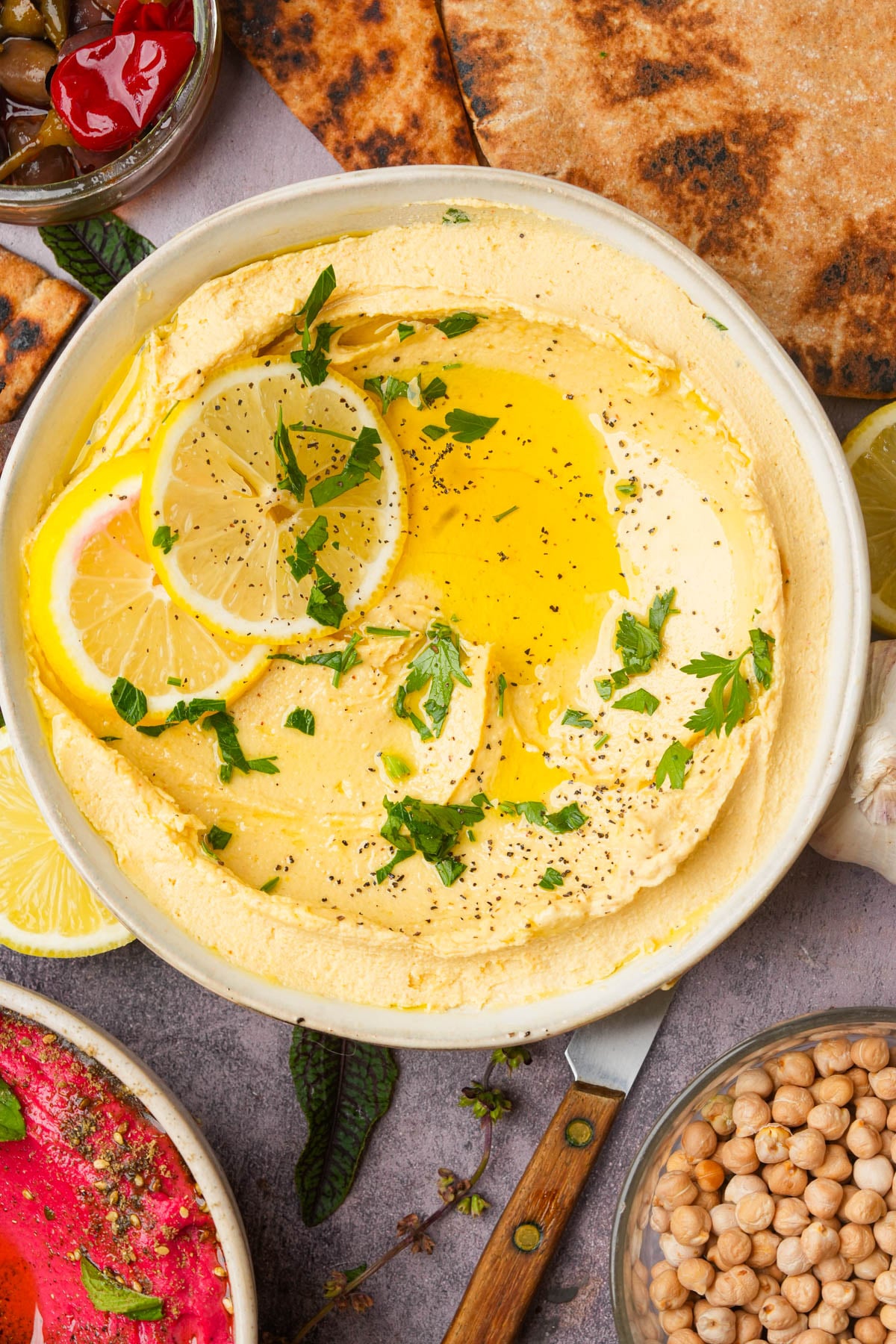 A bowl of creamy lemon hummus topped with olive oil, lemon slices, chopped parsley, and black pepper, surrounded by pita bread, chickpeas, garlic, olives, and a small bowl of pink dip.