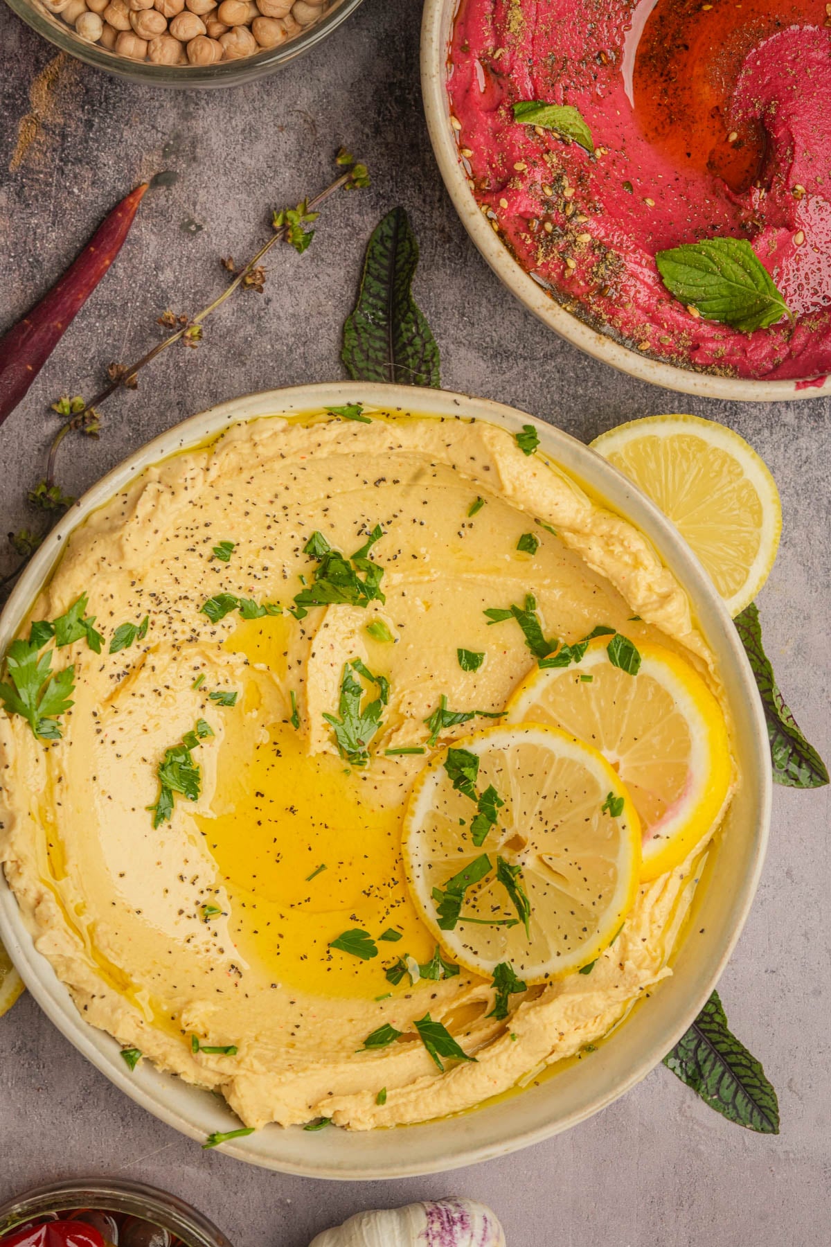 A bowl of creamy lemon hummus garnished with olive oil, black pepper, chopped parsley, and lemon slices, with a bowl of pink beet hummus and ingredients like chickpeas and herbs visible nearby.