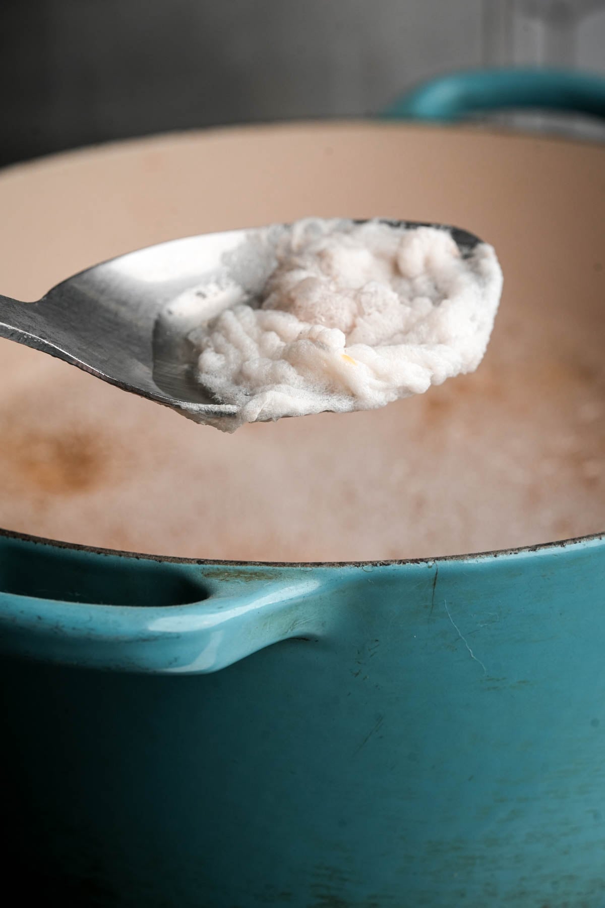 A metal spoon holds white scum skimmed from the surface of boiling liquid in a large blue pot.