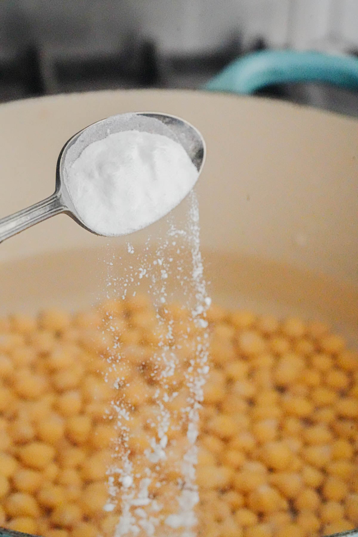 A spoonful of white baking soda is being sprinkled into a pot of boiling chickpeas.