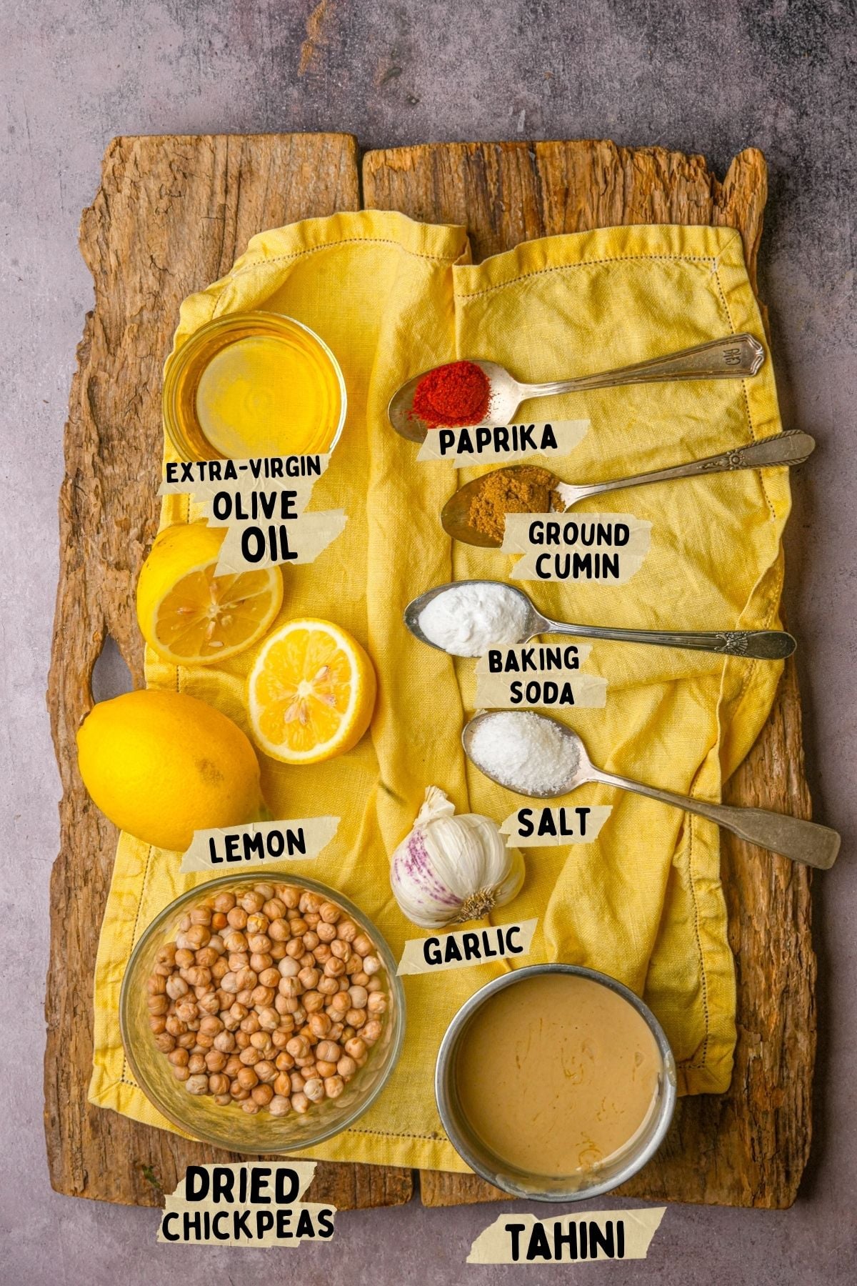 A rustic wooden board holds a yellow cloth with ingredients for a lemon hummus recipe: extra-virgin olive oil, paprika, ground cumin, baking soda, salt, lemon, garlic, dried chickpeas, and tahini in small bowls and spoons, each labeled.