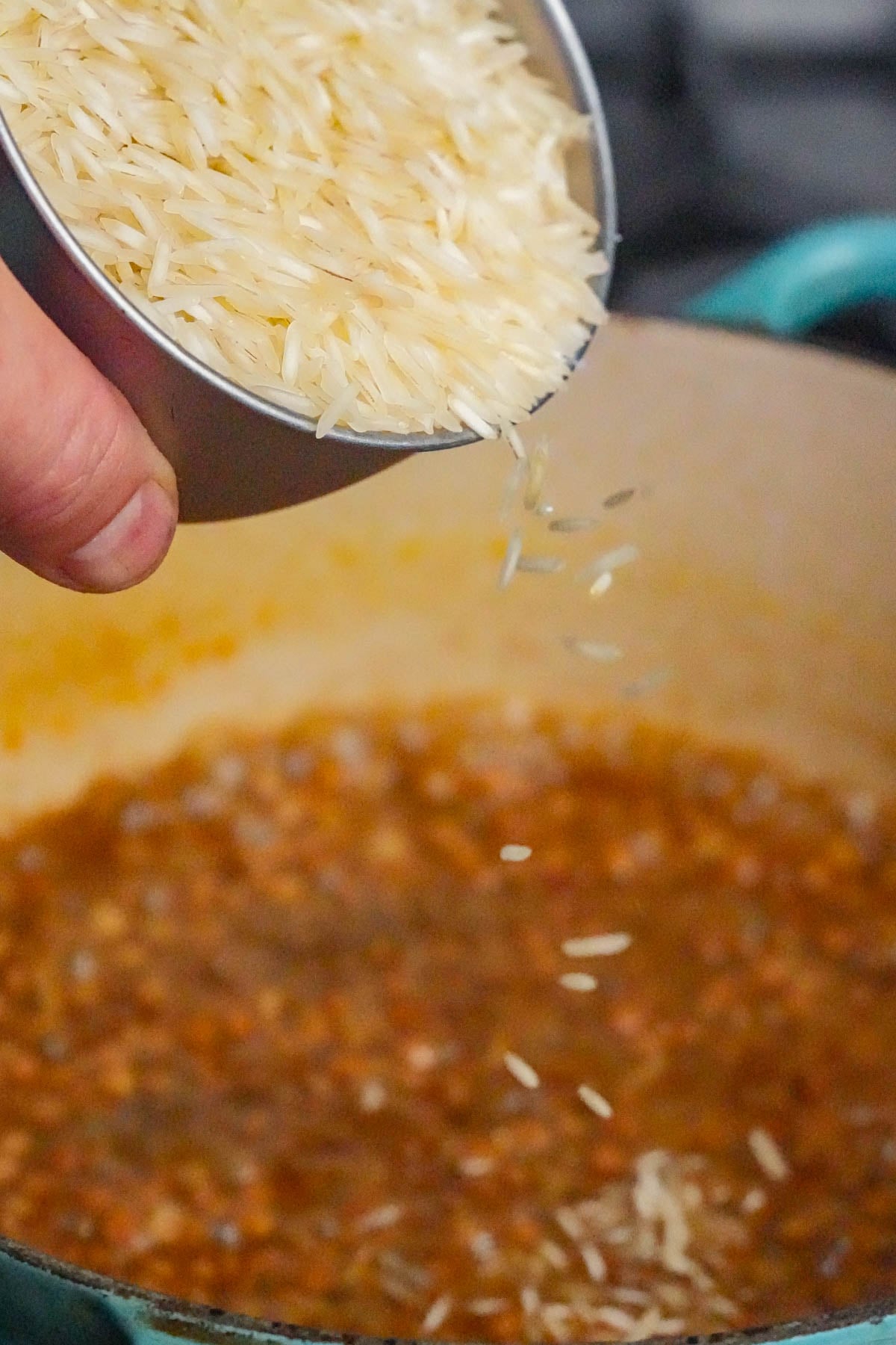 A hand pours uncooked basmati rice from a metal cup into a pot filled with a simmering, seasoned based for mujadara.
