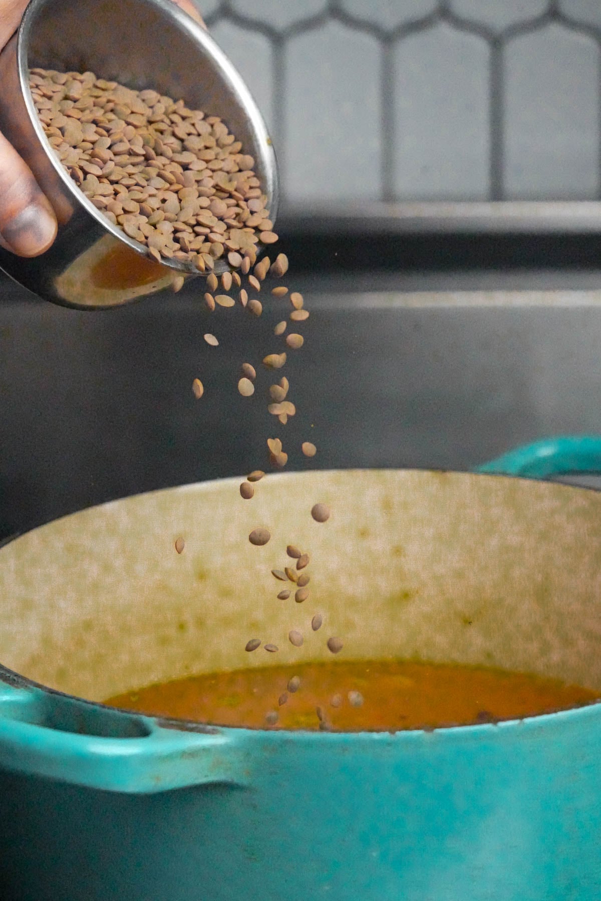 A hand pours brown lentils from a metal cup into a teal pot containing simmering bade for mujadara.