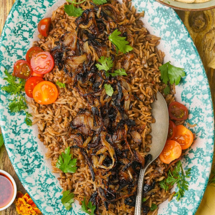 A platter of Mujadara-rice topped with caramelized onions, garnished with parsley and halved cherry tomatoes, with a serving spoon on the side.