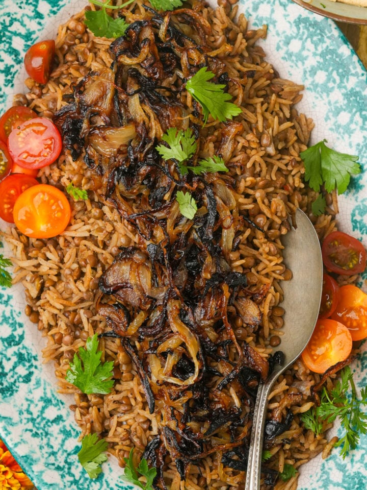 A platter of Mujadara—rice topped with caramelized onions, garnished with parsley and halved cherry tomatoes, with a serving spoon on the side.