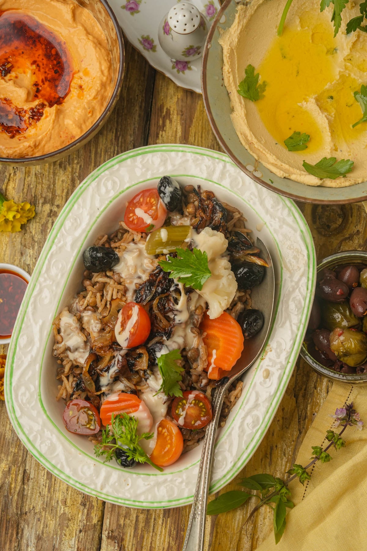 A bowl of mujadara with olives, cherry tomatoes, parsley, and sauce sits on a rustic wooden table, surrounded by hummus, dip, and more olives.