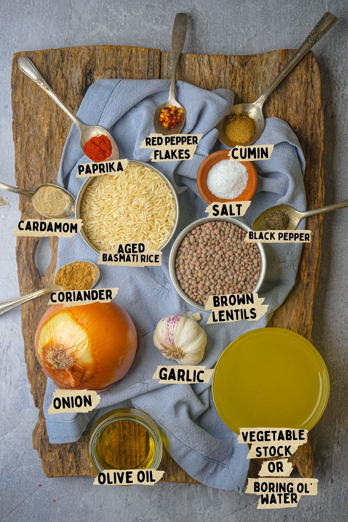Labeled ingredients for a mujadara recipe: aged basmati rice, brown lentils, onion, garlic, olive oil, vegetable stock, salt, black pepper, cardamom, coriander, cumin, paprika, and red pepper flakes on a board with measuring spoons.