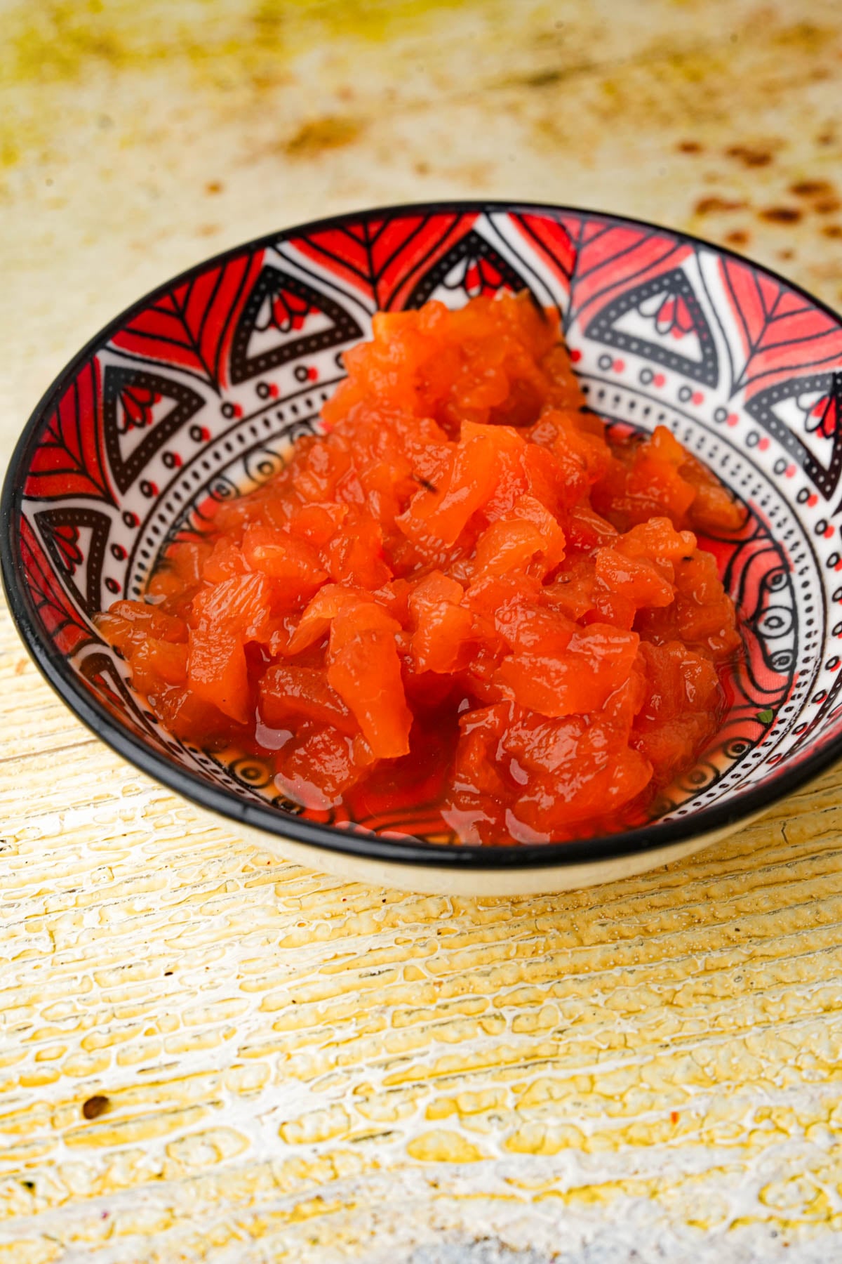 A decorative bowl with a red and black geometric pattern holds a serving of minced roasted red peppers placed on a rustic yellow surface.