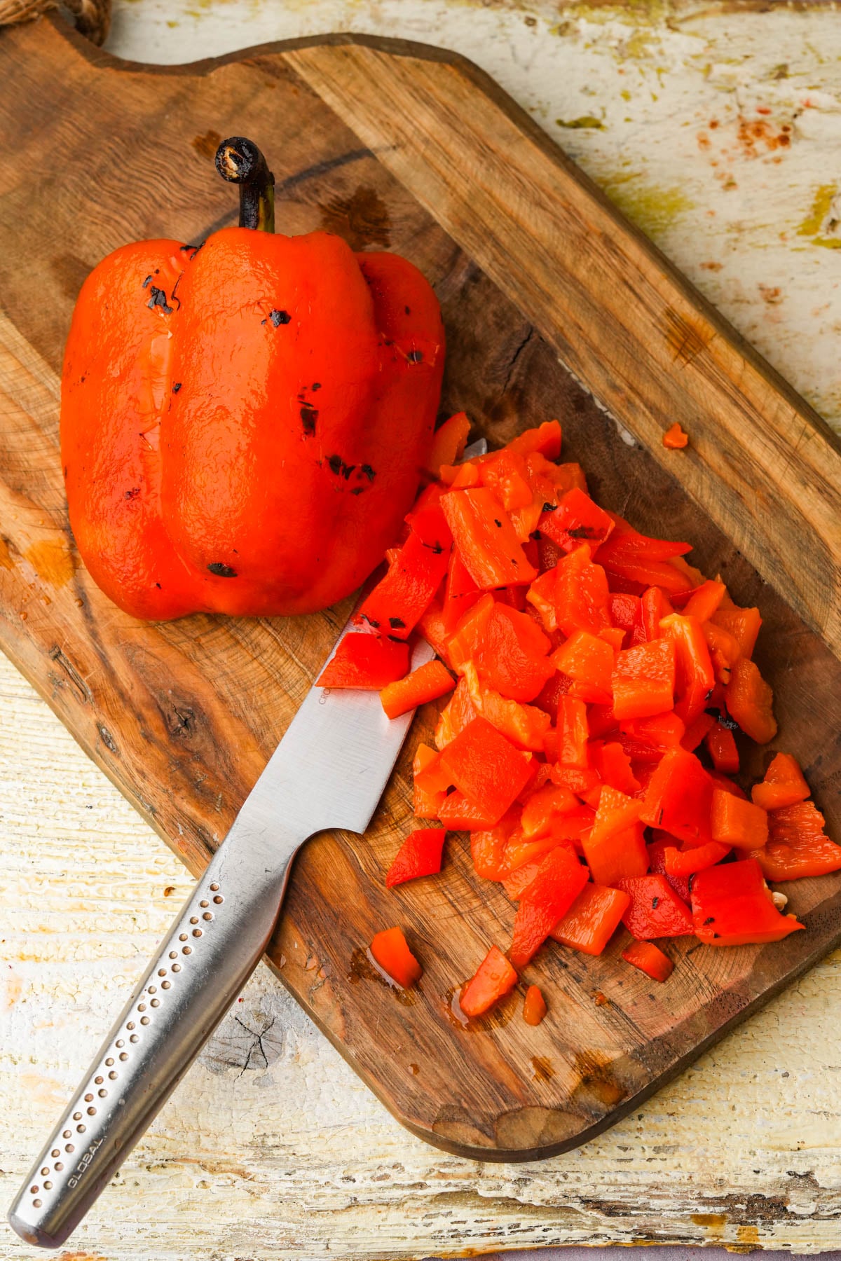 A partially chopped roasted red bell pepper and a pile of diced pepper pieces rest on a wooden cutting board beside a knife.