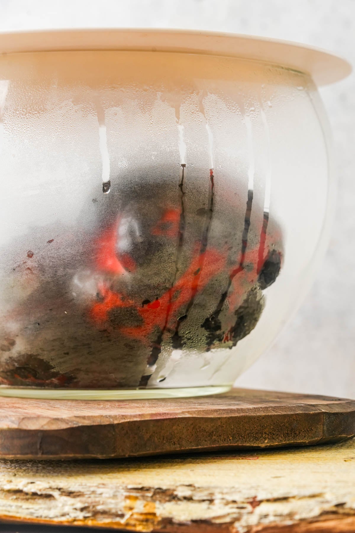 A close-up of a glass bowl covered with a plate, showing condensation and roasted red peppers inside on a wooden surface.