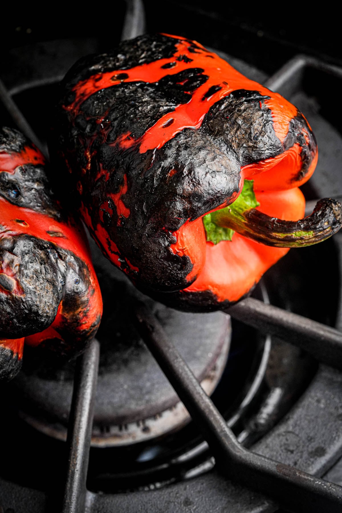 A red bell pepper with blackened, charred skin rests on a gas stove burner, being roasted over an open flame. Another partially visible pepper is also roasting beside it.