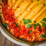 Roasted red pepper hummus topped with chopped parsley, red chili flakes, and a drizzle of red sauce, served in a rustic ceramic bowl on a wooden surface.