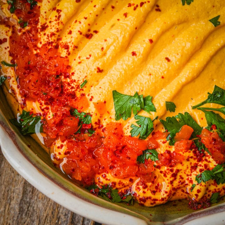 Roasted red pepper hummus topped with chopped parsley, red chili flakes, and a drizzle of red sauce, served in a rustic ceramic bowl on a wooden surface.