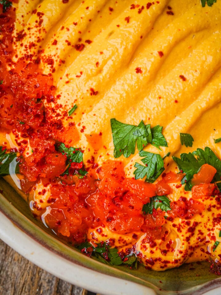 Roasted red pepper hummus topped with chopped parsley, red chili flakes, and a drizzle of red sauce, served in a rustic ceramic bowl on a wooden surface.