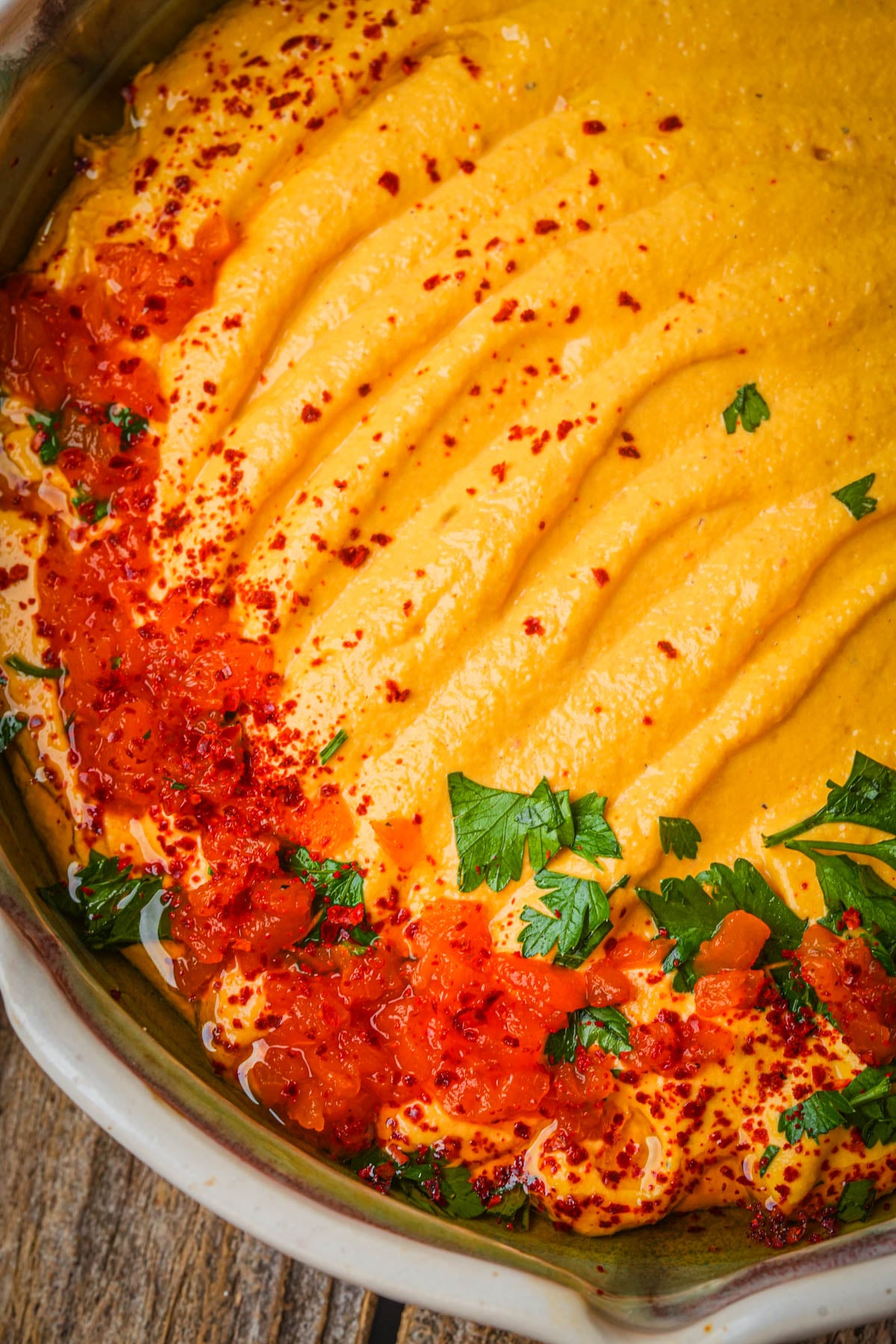 A close-up of creamy roasted red pepper hummus topped with red chili flakes, diced tomatoes, and fresh parsley, served in a shallow bowl on a wooden surface.