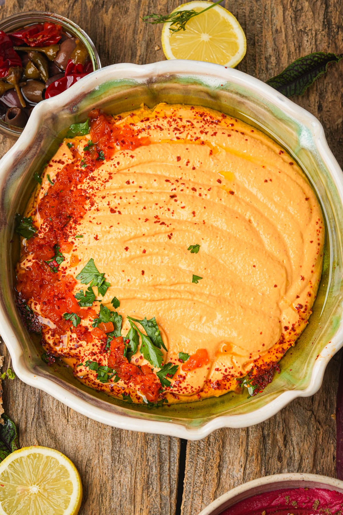 A bowl of creamy roasted red pepper hummus topped with olive oil, chopped herbs, red spices, and tomato relish, surrounded by lemon slices, olives, and herbs on a rustic wooden table.