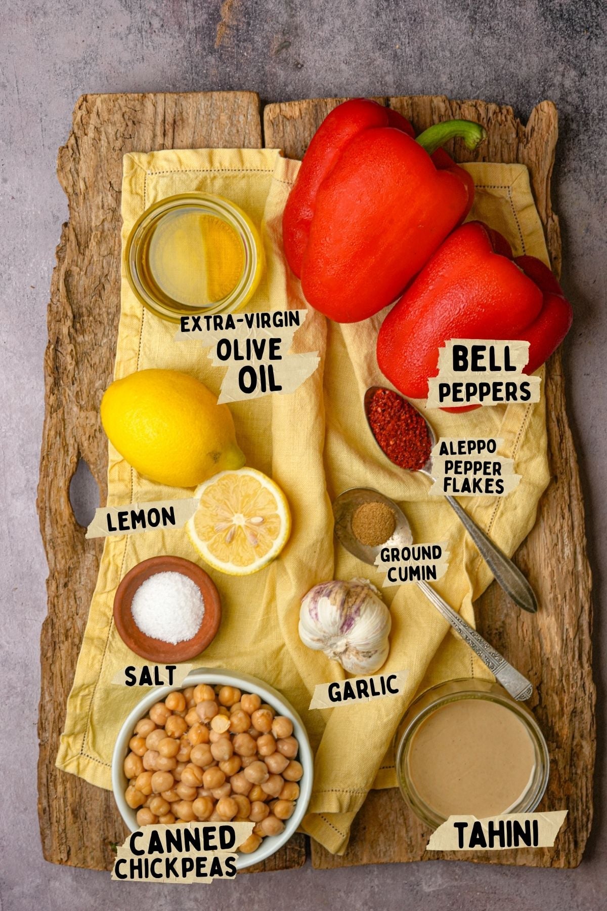 A wooden board with a yellow cloth displays labeled ingredients for a roasted red pepper hummus recipe: extra-virgin olive oil, two red bell peppers, lemon, Aleppo pepper flakes, ground cumin, salt, garlic, canned chickpeas, and tahini.