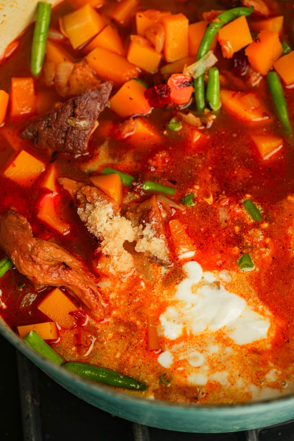 A pot of chipotle butternut squash soup with chunks of seitan, diced carrots, green beans, and a dollop of coconut milk in a rich, reddish broth.