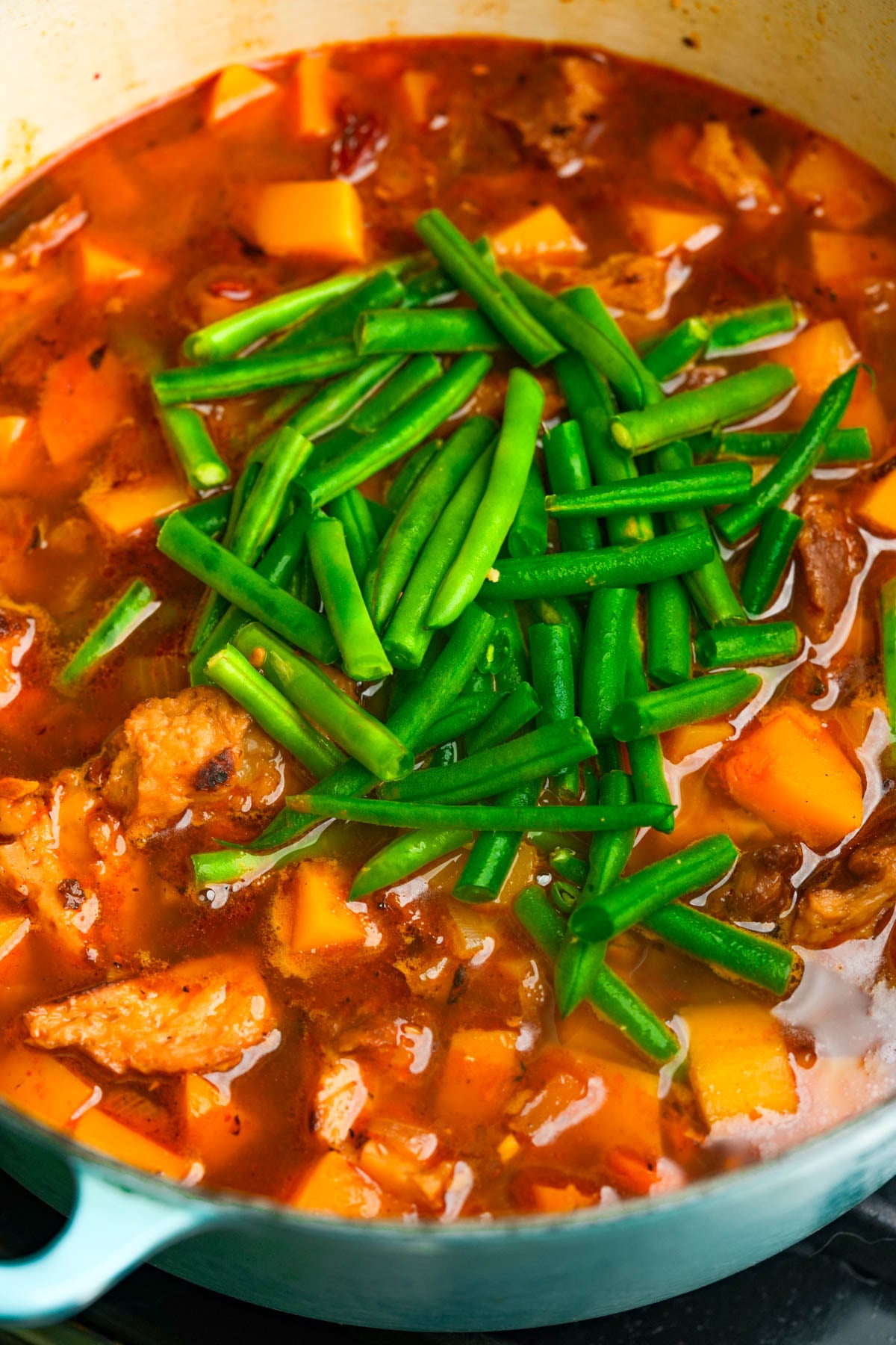 A pot of chipotle butternut squash soup with chunks of seitan, orange vegetables, and fresh green beans on top in broth.