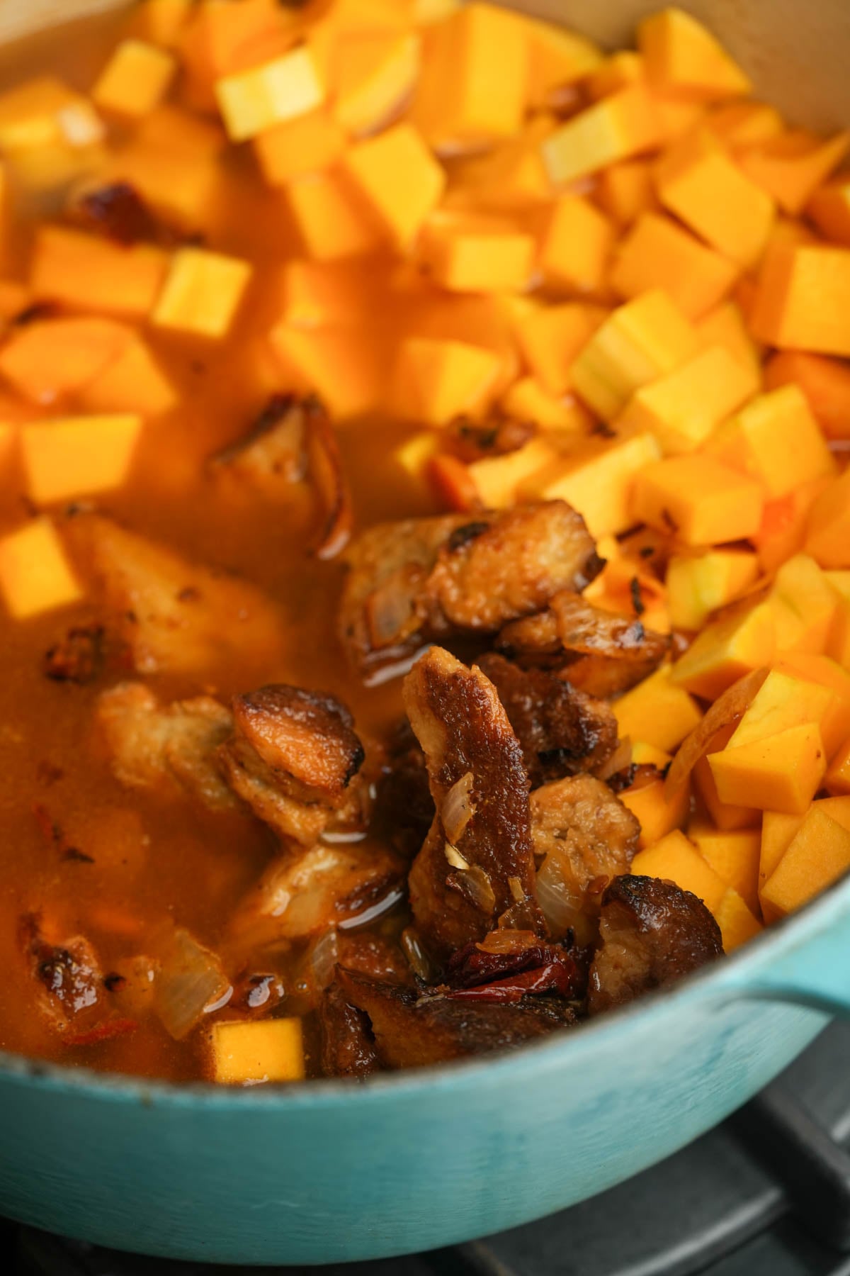 Diced butternut squash and browned seitan pieces simmer in a rich, orange broth inside a turquoise pot on a stovetop.