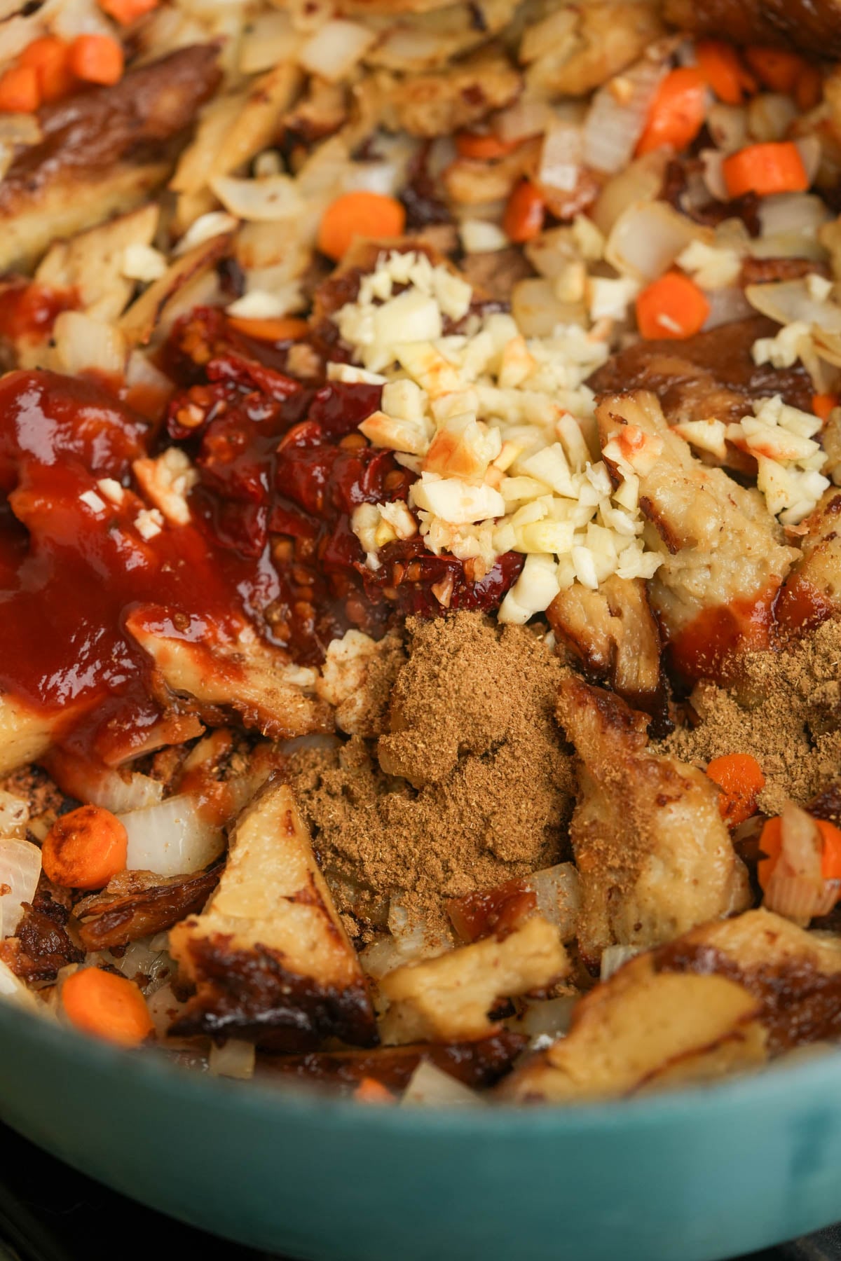 A skillet filled with sautéed onions, carrots, chopped garlic, shredded plant-based meat, chili, ground spices, and a dollop of chopped chipotle peppers in adobo, all about to be mixed together.