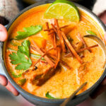 A bowl of creamy chipotle butternut squash soup garnished with cilantro, crispy strips, lime, and chunks of seitan and vegetables, with a spoon on the side.