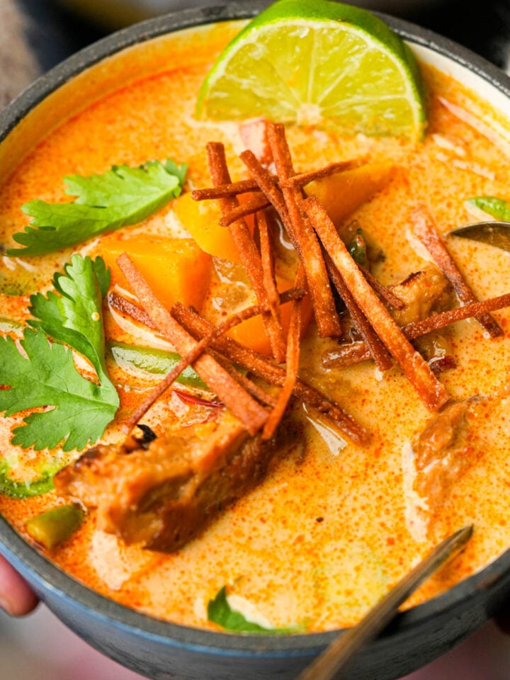 A bowl of creamy chipotle butternut squash soup garnished with cilantro, crispy strips, lime, and chunks of seitan and vegetables, with a spoon on the side.