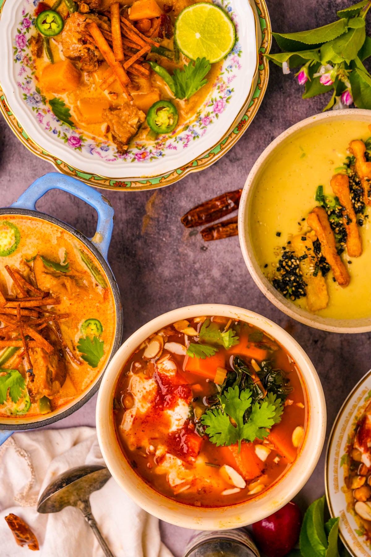 Four colorful bowls of chipotle butternut squash soup, japanese sweet potato soup, and Moroccan sweet potato soup, garnished with herbs and crispy toppings, arranged on a tabletop with scattered spices and a vintage spoon nearby.