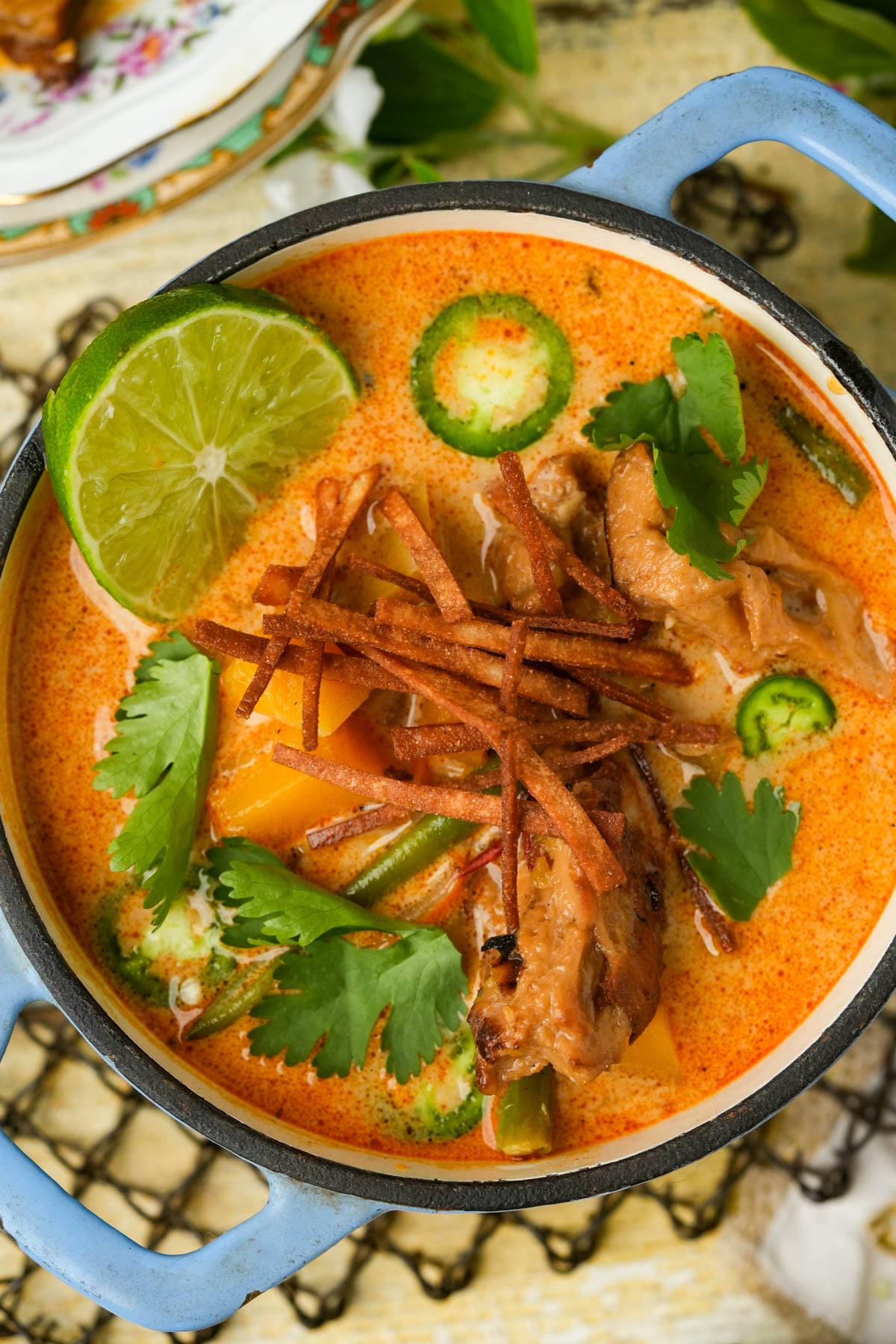 A bowl of creamy orange chipotle butternut squash soup with seitan, sliced jalapeños, cilantro, tortilla strips, vegetables, and a lime wedge, served in a blue pot.