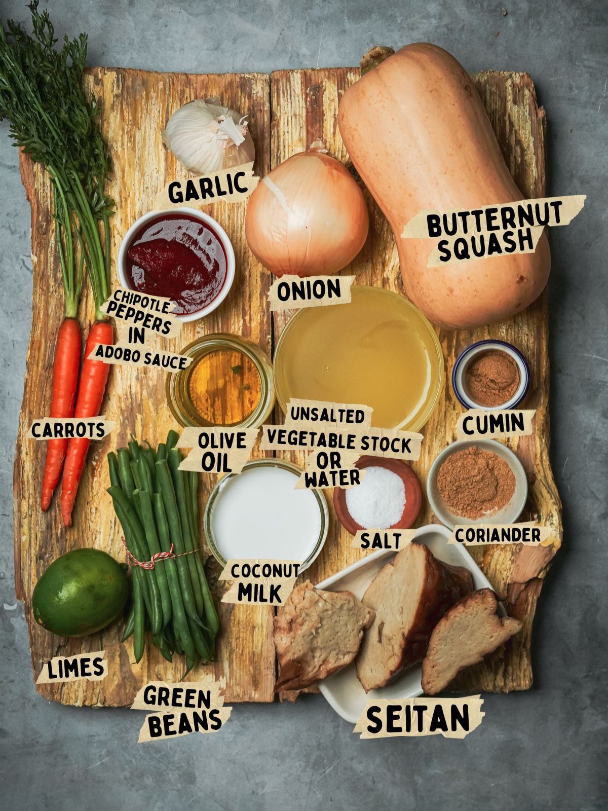 A top-down view of labeled ingredients for a chipotle butternut squash soup recipe on a wooden board, including butternut squash, seitan, carrots, green beans, limes, onion, garlic, chipotle peppers, olive oil, vegetable stock, coconut milk, cumin, coriander, and salt.